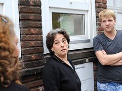 Drei Personen stehen vor einem Backsteinhaus mit weißen Fensterrahmen, zwei lehnen an der Wand, eine steht mit dem Rücken zur Kamera.