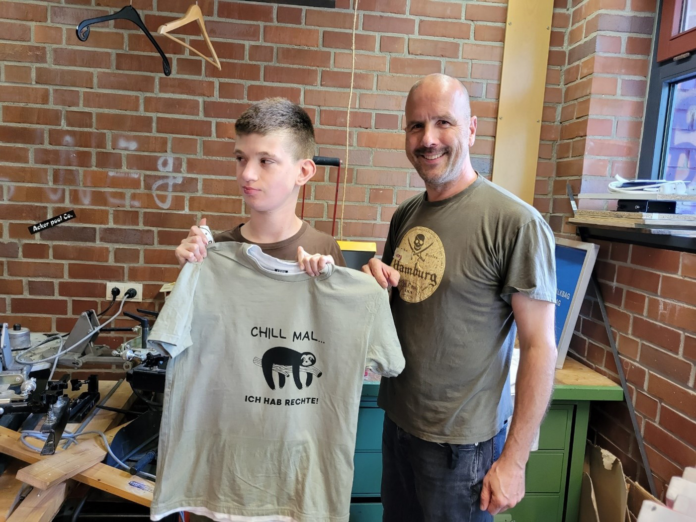 Workshop Wir machen Druck: jugendlicher Teilnehmer präsentiert sein bedrucktes T-Shirt mit der Botschaft: Chill mal, ich hab Rechte. Daneben ein Mitarbeiter