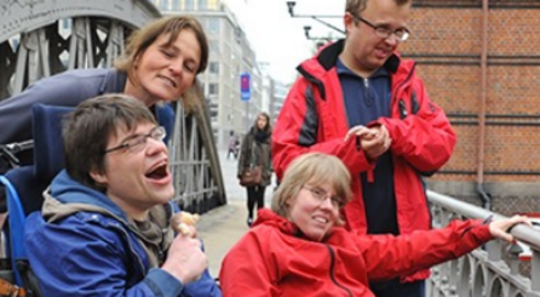 Zwei Menschen im Rollstuhl mit Begleitung auf einer Brücke