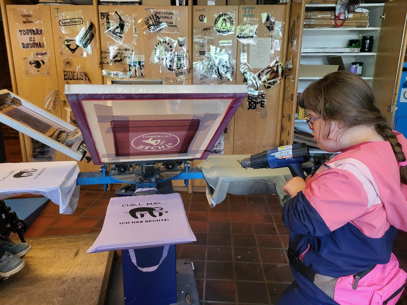 Workshop Wir machen Druck: jugendliche Teilnehmerin mit Heißluftfön vor einem eingespannten bedruckten T-Shirt