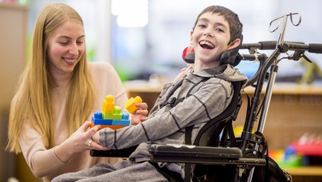 Frau reicht Spielzeug an Jungen im Rollstuhl.